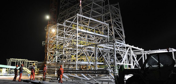 The structural framework of Soyuz’ mobile service gantry is flooded with light during nighttime work at the Spaceport (photo above). In the photo below, the four main support arms that will hold a Soyuz vehicle in position over the launch pad are visible in the foreground, while the illuminated mobile gantry is highlighted in its parked position 80 meters away. The structural framework of Soyuz’ mobile service gantry is flooded with light during nighttime work at the Spaceport (photo above). In the photo below, the four main support arms that will hold a Soyuz vehicle in position over the launch pad are visible in the foreground, while the illuminated mobile gantry is highlighted in its parked position 80 meters away.