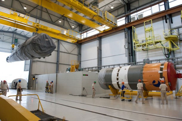 Suspended from the system of overhead mobile bridge cranes, the strap-on booster is moved into the Launcher Integration Building. Soyuz utilizes four of the strap-on boosters, which are clustered around the launcher’s Block A core stage. Also visible to the right in this photo is the Soyuz’ Block I third stage, which is resting on a horizontal jig. Suspended from the system of overhead mobile bridge cranes, the strap-on booster is moved into the Launcher Integration Building. Soyuz utilizes four of the strap-on boosters, which are clustered around the launcher’s Block A core stage. Also visible to the right in this photo is the Soyuz’ Block I third stage, which is resting on a horizontal jig.