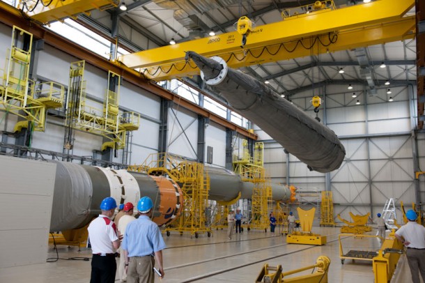 This image shows three main elements of the Soyuz launcher during its assembly process. The first stage strap-on booster is readied to be lowered onto its jig, while the Block I third stage is visible in the left foreground, and the Block A core second stage can be seen in the background. This image shows three main elements of the Soyuz launcher during its assembly process. The first stage strap-on booster is readied to be lowered onto its jig, while the Block I third stage is visible in the left foreground, and the Block A core second stage can be seen in the background.