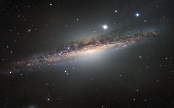 This colourful image from ESO’s Very Large Telescope shows NGC 1055 in the constellation of Cetus (The Sea Monster). This large galaxy is thought to be up to 15 percent larger in diameter than the Milky Way. NGC 1055 appears to lack the whirling arms characteristic of a spiral, as it is seen edge-on. However, it displays odd twists in its structure that were probably caused by an interaction with a large neighbouring galaxy. Credit: ESO This colourful image from ESO’s Very Large Telescope shows NGC 1055 in the constellation of Cetus (The Sea Monster). This large galaxy is thought to be up to 15 percent larger in diameter than the Milky Way. NGC 1055 appears to lack the whirling arms characteristic of a spiral, as it is seen edge-on. However, it displays odd twists in its structure that were probably caused by an interaction with a large neighbouring galaxy. Credit: ESO