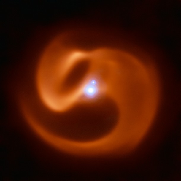 The reddish pinwheel in this image is data from the VISIR instrument on ESO’s Very Large Telescope (VLT), and shows the spectacular plumes of dust surrounding Apep. The blue sources at the centre of the image are a triple star system — which consists of a binary star system and a companion single star bound together by gravity. Though only two star-like objects are visible in the image, the lower source is in fact an unresolved binary Wolf-Rayet star. The triple star system was captured by the NACOadaptive optics instrument on the VLT. Credit: ESO/Callingham et al. The reddish pinwheel in this image is data from the VISIR instrument on ESO’s Very Large Telescope (VLT), and shows the spectacular plumes of dust surrounding Apep. The blue sources at the centre of the image are a triple star system — which consists of a binary star system and a companion single star bound together by gravity. Though only two star-like objects are visible in the image, the lower source is in fact an unresolved binary Wolf-Rayet star. The triple star system was captured by the NACOadaptive optics instrument on the VLT. Credit: ESO/Callingham et al.
